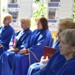 Grupa osób w niebieskich szatach siedzi na krzesłach podczas uroczystości na świeżym powietrzu.