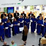 Zespół wokalny w niebieskich szatach śpiewa na scenie w bibliotece, obserwując dyrygentkę.