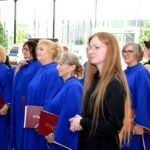 Grupa kobiet w niebieskich togach i czarnych ubraniach stoi razem, trzymając czerwone teczki, na tle okiennic i tablic informacyjnych.