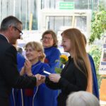 Mężczyzna wręcza nagrodę młodej kobiecie w czarnej sukience, otoczeni przez grupę kobiet w kolorowych szatach.
