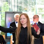 Kobieta o rudych włosach w okularach i czarnej koszuli wystawia rękę podczas uroczystości, otoczona mężczyznami w garniturach.