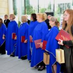 Grupa osób w błękitnych szatach trzymających czerwone teczki stoi w kolejce w jasnym pomieszczeniu.