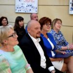 Grupa osób siedzi na krzesłach podczas wydarzenia, prawdopodobnie koncertu chóralnego.