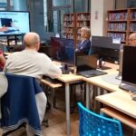 Uczestnicy szkolenia siedzą przy komputerach w sali wyposażonej w regały z książkami.