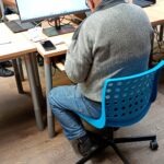 Mężczyzna siedzi przed komputerem na stanowisku z niebieskim krzesłem.