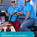 Dwie pracownice biblioteki prezentują mobilną wypożyczalnię książek zamontowaną na rowerze podczas Nocy Bibliotek.