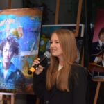 Młoda kobieta trzyma mikrofon, prezentując portret inspirowany zespołem The Beatles w trakcie wystawy malarstwa.