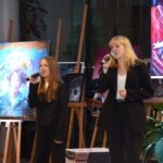 Dwie kobiety śpiewają do mikrofonów w towarzystwie kilku sztalug z obrazami.