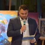 Mężczyzna w garniturze trzyma mikrofon i notatki, prezentując nagrody podczas wydarzenia związanego z wystawą malarstwa.