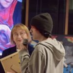 Młodzieżowy artysta z mikrofonem rozmawia z kobietą w okularach podczas wydarzenia w bibliotece.