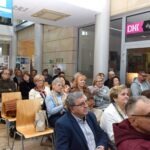 Publiczność zgromadzona jest w sali, prawdopodobnie podczas wydarzenia związanego z wystawą malarstwa.