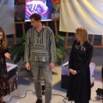 Mężczyzna w charakterystycznej koszuli stoi na scenie podczas wydarzenia związanego z wystawą malarstwa.