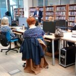 Uczestnicy warsztatów siedzą przy stolikach z komputerami w pomieszczeniu bibliotecznym, a w tle widoczne są regały z książkami i mężczyzna w garniturze.