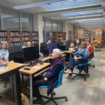 Uczestnicy warsztatów siedzą przy komputerach w jasnym pomieszczeniu bibliotecznym, korzystając z dostępnych stanowisk pracy.