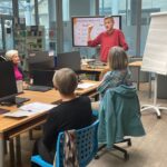 Prowadzący prezentację wskazuje na wykres wyświetlany na ekranie, a uczestnicy warsztatów siedzą przy stolikach z laptopami i dokumentami.