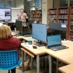 Na sali szkoleniowej uczestnicy warsztatów siedzą przed komputerami, a w tle widoczne są regały z książkami.