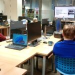 Na biurku znajdują się laptopy, monitory oraz smartfon, a w tle widoczne są inne stanowiska pracy.