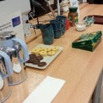 Na stole znajdują się filiżanki, talerz z ciasteczkami, czajnik i paczka herbaty.