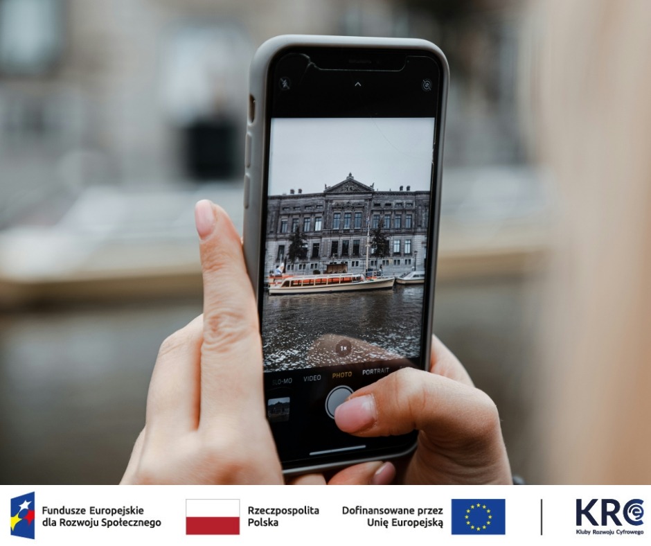 Na ekranie smartfona widoczny jest czarno-biały kadr przedstawiający budynek i łódź pływającą po wodzie.