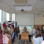 Uczniowie słuchają prezentacji przed ekranem projekcyjnym w szkolnej bibliotece.