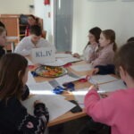 Uczniowie siedzą przy stole, pracując nad zadaniami z kaligrafii i papierniczych materiałów.