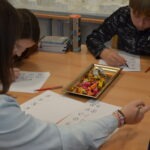 Uczniowie siedzą przy stole i piszą, a na blacie leży metalowa tacka z kredkami.