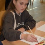 Dziewczynka siedzi przy stole i pisze ołówkiem do ćwiczeń kaligraficznych.