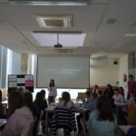 Uczeń prezentuje wystąpienie przed publicznością siedzącą w sali bibliotecznej z projekcją na ekranie.