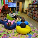 Dzieci siedzą na workach sensorycznych rozproszonych na kolorowej macie podczas zajęć w bibliotece.