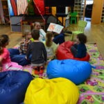 Dzieci siedzą na workach sensorycznych podczas czytania książki przez prowadzącą zajęcia.