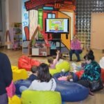 Dzieci siedzą na miękkich siedziskach, skupione na projekcji edukacyjnej prezentacji na ekranie telewizora.