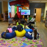 Dzieci siedzą na workach do siedzenia i oglądają film wyświetlany na ekranie podczas spotkania Dyskusyjnego Klubu Książki.