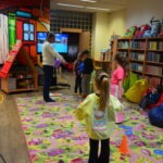 Dzieci uczestniczą w zajęciach Dyskusyjnego Klubu Książki, otoczone książkami i zabawkami w kolorowym pomieszczeniu.