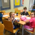 Dzieci siedzą przy stole w sali lekcyjnej, skupione na pracy plastycznej z plasteliną i cekinami.