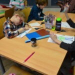 Dzieci siedzą przy stole i pracują nad projektami plastycznymi, wykorzystując plastelinę i cekiny.