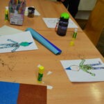 Na stole leżą kartki z rysunkami pępków oraz materiały plastyczne, takie jak kredki, klej i kolorowy papier.