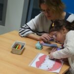 Dziecko maluje kredkami pod nadzorem dorosłej osoby przy stole, obok leżą pojemniczki z materiałami plastycznymi.