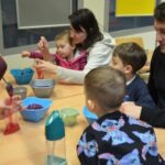 Dzieci pod opieką dorosłych uczestniczą w zabawie edukacyjnej, korzystając z misek wypełnionych kolorowym kisielem.