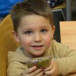 Chłopiec trzyma w ręku miseczkę z jedzeniem, uśmiechając się do kamery.
