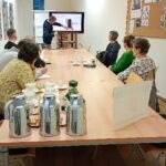 Uczestnicy warsztatów siedzą przy długim stole w sali konferencyjnej, podczas gdy prowadzący prezentuje coś na ekranie telewizora.