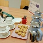 Na stole znajdują się filiżanki z porcelany, talerzyki, koszyk z ciasteczkami i miedziana kawiarka.