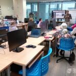 Uczestnicy warsztatów siedzą przy stolikach z komputerami, słuchając wykładowcy prezentującego materiał na ekranie.