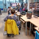 Uczestnik warsztatów siedzi przy biurku z komputerem, a w tle widoczne są inne osoby i ekran telewizora.