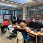 Uczestnicy warsztatów siedzą przy komputerach w bibliotece, a na ekranie wyświetlana jest prezentacja.