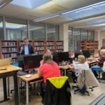 Uczestnicy warsztatów słuchają wykładowcy w pomieszczeniu z regałami pełnymi książek i komputerami.