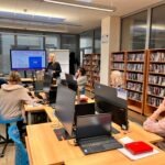 Uczestnicy warsztatów słuchają prezentacji przy ekranie w bibliotece otoczonej regałami z książkami.