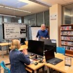 Uczestnik warsztatów siedzi przy biurku z laptopem, a w tle edukator prezentuje coś na tablicy.