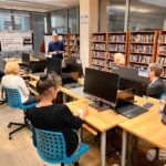 Uczestnicy warsztatów siedzą przy komputerach, a edukator prezentuje informacje na tablicy.