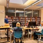 Uczestnicy warsztatów słuchają wykładowcy w pomieszczeniu z regałami pełnymi książek.