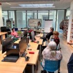 Uczestnicy warsztatów siedzą przy stolikach z komputerami, a w tle widoczne jest tablicę i okna.